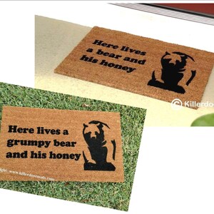 May include: A brown coir doormat with the text "Here lives a grumpy bear and his honey" and a black silhouette of a bear. The text "Copyright: www.killerdoormats.com" is at the bottom of the mat.