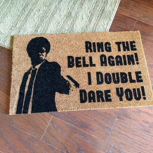 May include: A brown doormat with the text "Ring the bell again! I double dare you!" in black lettering. A silhouette of a man in a suit is on the left side of the doormat.
