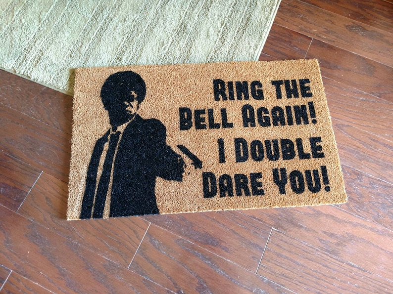 May include: A brown doormat with the text "Ring the bell again! I double dare you!" in black lettering. A silhouette of a man in a suit is on the left side of the doormat.