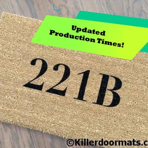 May include: A brown coir doormat with the number "221B" printed in black. The doormat has a red and white striped design in the upper right corner. The text "Updated Production Times!" is visible in a green and yellow banner.