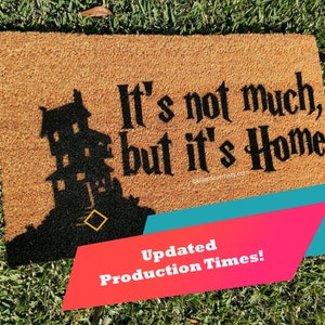 May include: A brown coir doormat with the text "It's not much, but it's Home" in black lettering. A black silhouette of a house is on the left side of the mat.