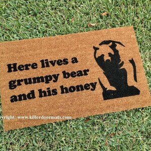 May include: A brown coir doormat with the text "Here lives a grumpy bear and his honey" in black lettering. A black silhouette of a bear is on the right side of the mat.