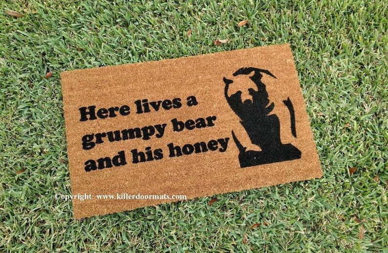 May include: A brown coir doormat with the text "Here lives a grumpy bear and his honey" in black lettering. A black silhouette of a bear is on the right side of the mat.