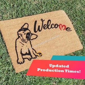 May include: A brown coir doormat with the text "Welcome" in black with a red heart in the "e". A black and white illustration of a French bulldog is on the left side of the mat.
