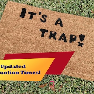 May include: A brown coir doormat with the text "IT'S A TRAP!" in black lettering. The doormat is on a green grassy surface.