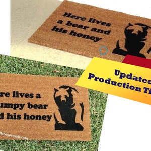 May include: A brown coir doormat with the text "Here lives a grumpy bear and his honey" in black lettering. A black silhouette of a bear is on the right side of the mat.