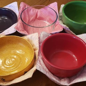 May include: Five bowls of different colors and materials are displayed on a wooden table. Each bowl is nestled in a fabric bowl cozy with a floral pattern. The bowl cozies are pink, white, and green.