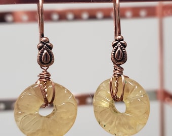 May include: A pair of earrings with a copper wire wrapped around a carved yellow stone. The stone is shaped like a ring with a flower design. The earrings have a copper hook with a decorative design.