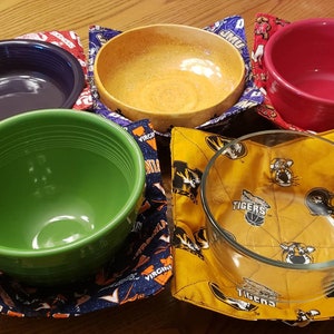 May include: Six bowls of different colors and materials are displayed on fabric squares with various college logos. The bowls include a green ceramic bowl, a purple ceramic bowl, a yellow ceramic bowl, a red ceramic bowl, and two glass bowls. The fabric squares feature logos for the University of Virginia, Oklahoma Sooners, Missouri Tigers, and James Madison University.