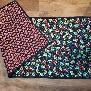 May include: Two black placemats with a black border. One placemat has a red and pink heart pattern. The other placemat has a green, red, and yellow mitten pattern.