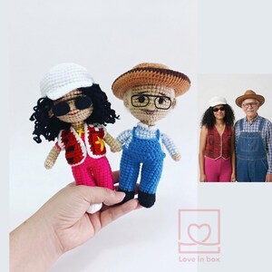 May include: Two crocheted dolls, one in a red vest and pink trousers, the other in blue dungarees. Both dolls wear hats and glasses. A smaller image shows the dolls' inspiration. The dolls are held in a hand.