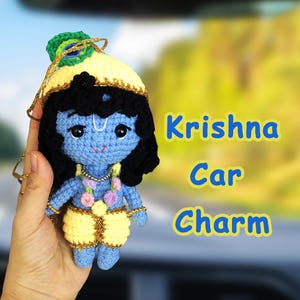 Könnte beinhalten: Eine gehäkelte Krishna-Puppe mit blauer Haut, schwarzem Haar, einem gelben Kopfschmuck mit Pfauenfeder und gelber Hose. Die Puppe trägt eine Blumengirlande und hat eine goldene Schnur zum Aufhängen. Der Text Krishna Car Charm ist zu sehen.