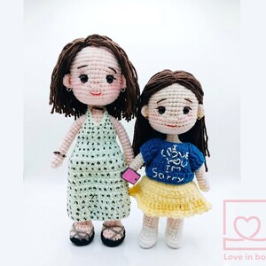 May include: Two handmade crochet dolls. One doll wears a light green halter dress with black polka dots, and the other wears a blue shirt with the text "I love you, I'm sorry" and a yellow skirt. Both dolls have brown hair and are holding a pink phone.