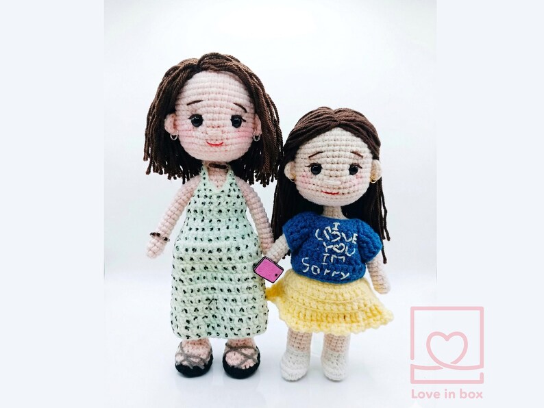 May include: Two handmade crochet dolls. One doll wears a light green halter dress with black polka dots, and the other wears a blue shirt with the text "I love you, I'm sorry" and a yellow skirt. Both dolls have brown hair and are holding a pink phone.