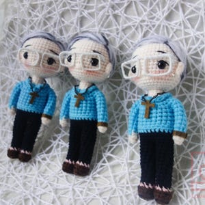 May include: Three crocheted dolls, each with grey hair, glasses, and a blue sweater featuring a cross pendant. The dolls are dressed in black trousers and brown shoes. They are displayed on a white, textured surface.