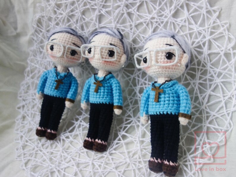May include: Three crocheted dolls, each with grey hair, glasses, and a blue sweater featuring a cross pendant. The dolls are dressed in black trousers and brown shoes. They are displayed on a white, textured surface.