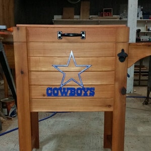May include: A wooden cooler with a blue star and the word "COWBOYS" on the front. The cooler has a black handle and a bottle opener on the side.