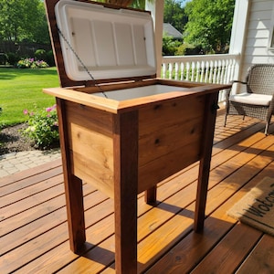 May include: A wooden cooler with a hinged lid, open to reveal a white interior. The cooler is constructed from light and dark brown wood and stands on four sturdy legs. The cooler is designed for outdoor use.