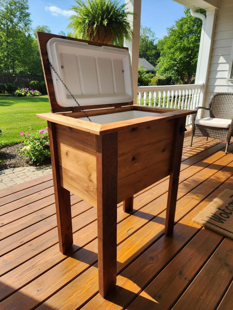 May include: A wooden cooler with a hinged lid, open to reveal a white interior. The cooler is constructed from light and dark brown wood and stands on four sturdy legs. The cooler is designed for outdoor use.