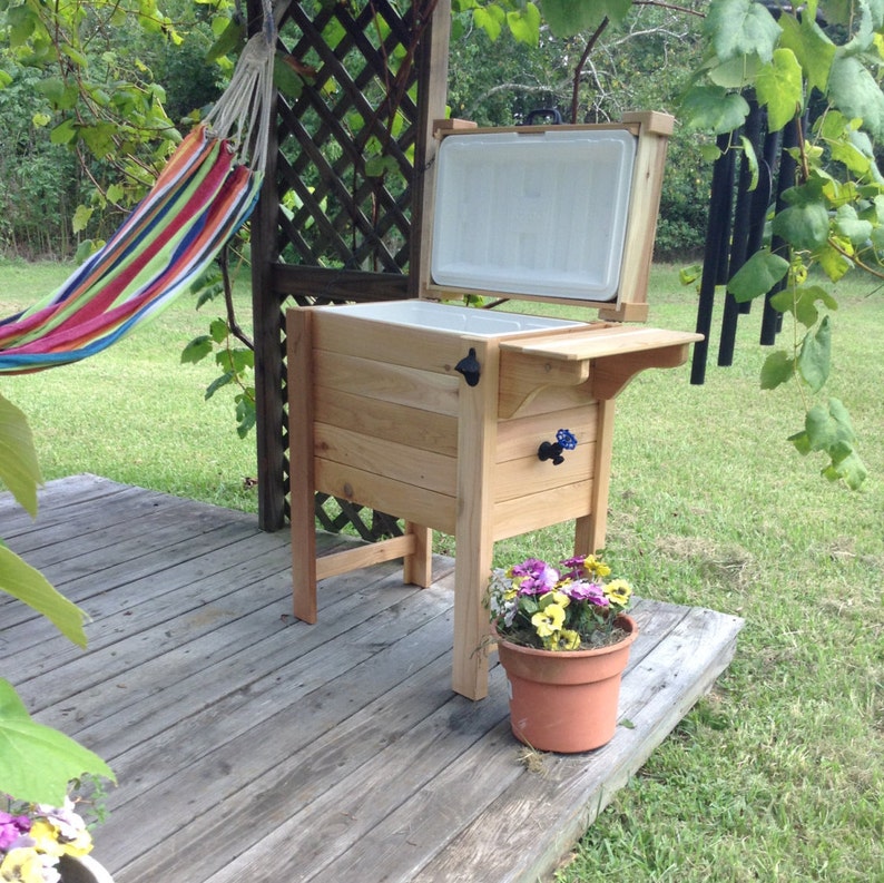 May include: A wooden cooler with a hinged lid, bottle opener, and spigot. The cooler is light brown and sits on a wooden deck. A flower pot with purple and yellow flowers is in front of the cooler. A hammock hangs in the background.
