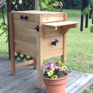 May include: A wooden outdoor cooler with a small side table and a bottle opener. The cooler has a black handle and a blue spigot. A terracotta pot with purple and yellow flowers sits in front of the cooler.
