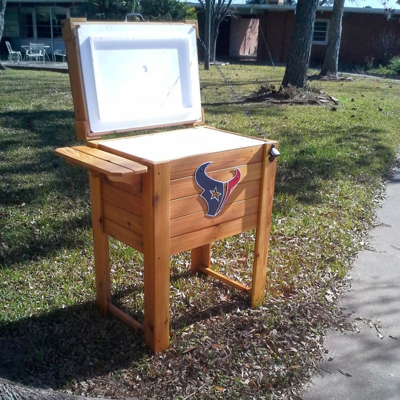 Puede incluir: Una nevera de madera con una tapa blanca y un logotipo de los Houston Texans en la parte delantera. La nevera est&aacute; sobre un soporte con un peque&ntilde;o estante en el lateral.