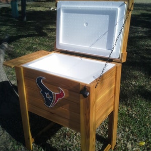 Puede incluir: Una nevera de madera con un revestimiento aislante blanco y un logotipo de los Houston Texans. La nevera est&aacute; abierta y tiene una cadena unida a la tapa.