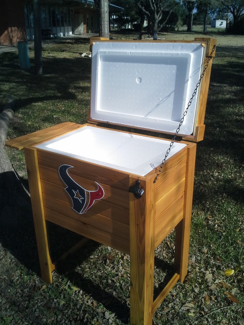 Puede incluir: Una nevera de madera con un revestimiento aislante blanco y un logotipo de los Houston Texans. La nevera est&aacute; abierta y tiene una cadena unida a la tapa.