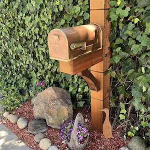 May include: A copper-colored metal mailbox with a brass-toned flag and the embossed text U.S. MAIL, mounted on a vertical wooden post. The mailbox features a curved top and a rectangular base, supported by a wooden bracket attached to the post.