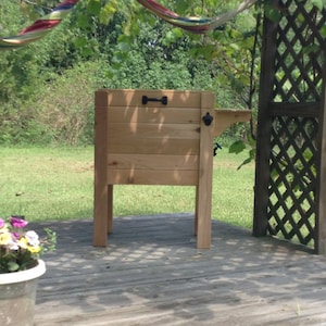May include: A wooden cooler with a built-in bottle opener and a handle. The cooler is made of light-colored wood and stands on four legs. It is set on a wooden deck, with plants and a hammock in the background.