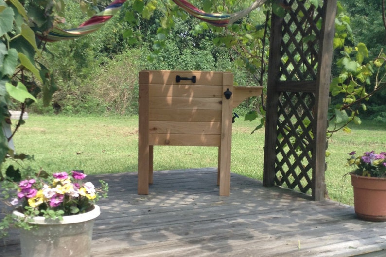 May include: A wooden cooler with a built-in bottle opener and a handle. The cooler is made of light-colored wood and stands on four legs. It is set on a wooden deck, with plants and a hammock in the background.