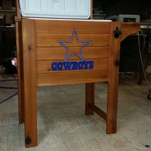 May include: A wooden cooler with a white lid and a blue star with the word "COWBOYS" underneath. The cooler is on a wooden stand with a bottle opener.