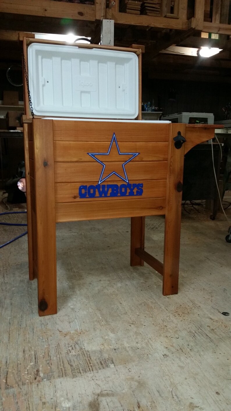 May include: A wooden cooler with a white lid and a blue star with the word "COWBOYS" underneath. The cooler is on a wooden stand with a bottle opener.