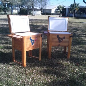 Puede incluir: Dos neveras de madera con las tapas abiertas, ambas con el logotipo de los Houston Texans. Las neveras est&aacute;n sobre una superficie cubierta de hierba.