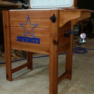 May include: A wooden cooler with a blue star and the word "COWBOYS" on the front. The cooler has a white lid that is open. The cooler is sitting on a wooden stand.