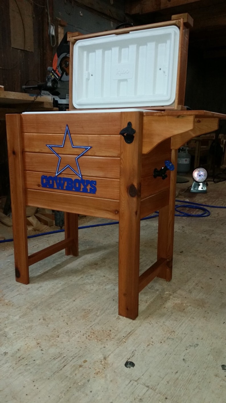 May include: A wooden cooler with a blue star and the word "COWBOYS" on the front. The cooler has a white lid that is open. The cooler is sitting on a wooden stand.