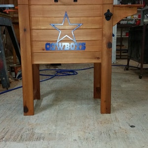 May include: A wooden cooler with a blue star and the word "COWBOYS" on the front. The cooler is on a stand with a bottle opener on the side.