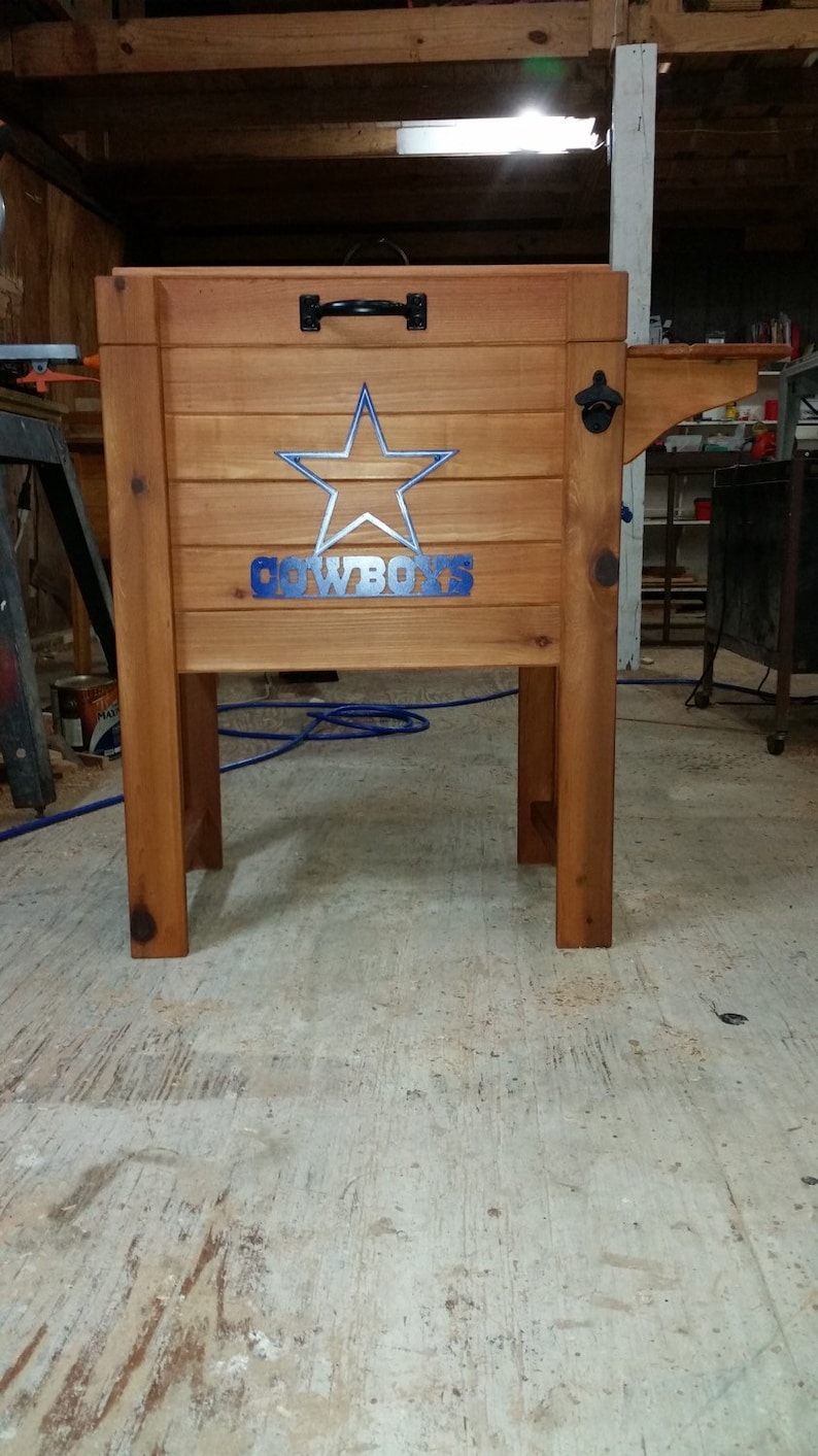 May include: A wooden cooler with a blue star and the word "COWBOYS" on the front. The cooler is on a stand with a bottle opener on the side.