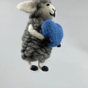 Puede incluir: Adorno de oveja gris hecho a mano sosteniendo un corazón azul. La oveja tiene orejas blancas y una cara blanca con ojos negros. Colgante de una cuerda de hilo natural. Una decoración encantadora y caprichosa.