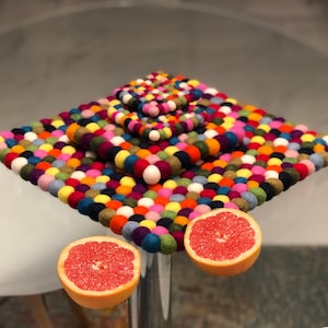 May include: A set of four colourful felt coasters with a round, bumpy texture. The coasters are stacked on top of each other, with the largest coaster on the bottom and the smallest coaster on top. The coasters are made of felt balls that are sewn together. The coasters are a variety of colours, including red, orange, yellow, green, blue, purple, and pink.