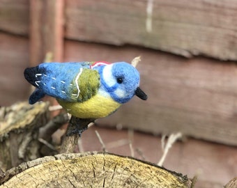 May include: A bluebird with a yellow belly and a white stripe on its head is perched on a branch of a tree stump. The bird is made of felt and has a stitched-on eye.