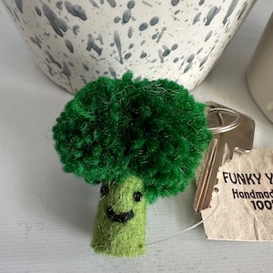 Puede incluir: Un llavero de brócoli de fieltro verde con una cara sonriente. El llavero está unido a un llavero plateado y una etiqueta que dice "FUNKY YARN Hecho a mano 100%"