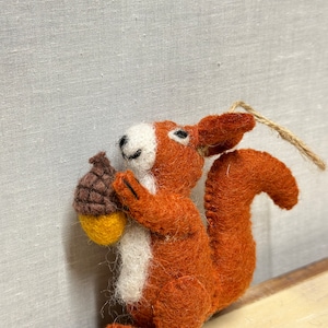 Könnte beinhalten: Handgefertigter Eichhörnchen-Ornament aus Filz in Orange-, Weiß- und Brauntönen, das eine Eichel hält. Das Eichhörnchen hat einen flauschigen Schwanz und ist an einer natürlichen Schnurschlaufe aufgehängt. Die Eichel ist gelb und braun.