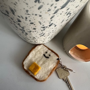 Puede incluir: Un llavero de fieltro con forma de rebanada de pan con una cara sonriente y un cuadrado amarillo de fieltro para la mantequilla. El llavero está unido a una llave plateada.