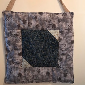 May include: A handmade tote bag with a gray and white leaf pattern. The center features a dark blue square with a delicate gold and white floral design. The bag has a beige strap and is made of quilted fabric.