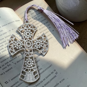 May include: A silver-colored, openwork cross-shaped bookmark with a lavender tassel. The bookmark is made of delicate, interwoven threads, and is resting on the pages of an open book.