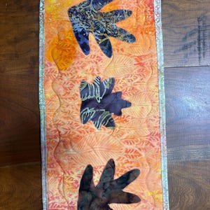 May include: A rectangular quilted wall hanging featuring an orange background with a subtle leaf pattern. Three dark, batik-style fabric leaf appliques are stitched vertically down the center. The piece is finished with a light green border.