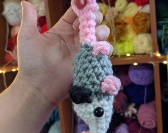 May include: A handmade crochet opossum toy in gray, white, black, and pink. The opossum has a pink tail and small pink feet. The toy is held in a hand, with a shelf of yarn in the background.