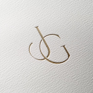 May include: A gold-colored monogram featuring the intertwined letters "J" and "G" is embossed on a textured, off-white surface. The elegant design suggests a logo or branding element, possibly for a business or personal project.