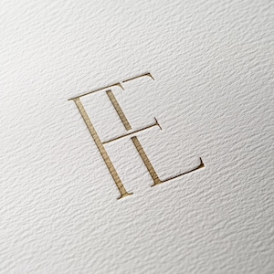 May include: A gold-colored monogram of the letter "E" with a stylized design.